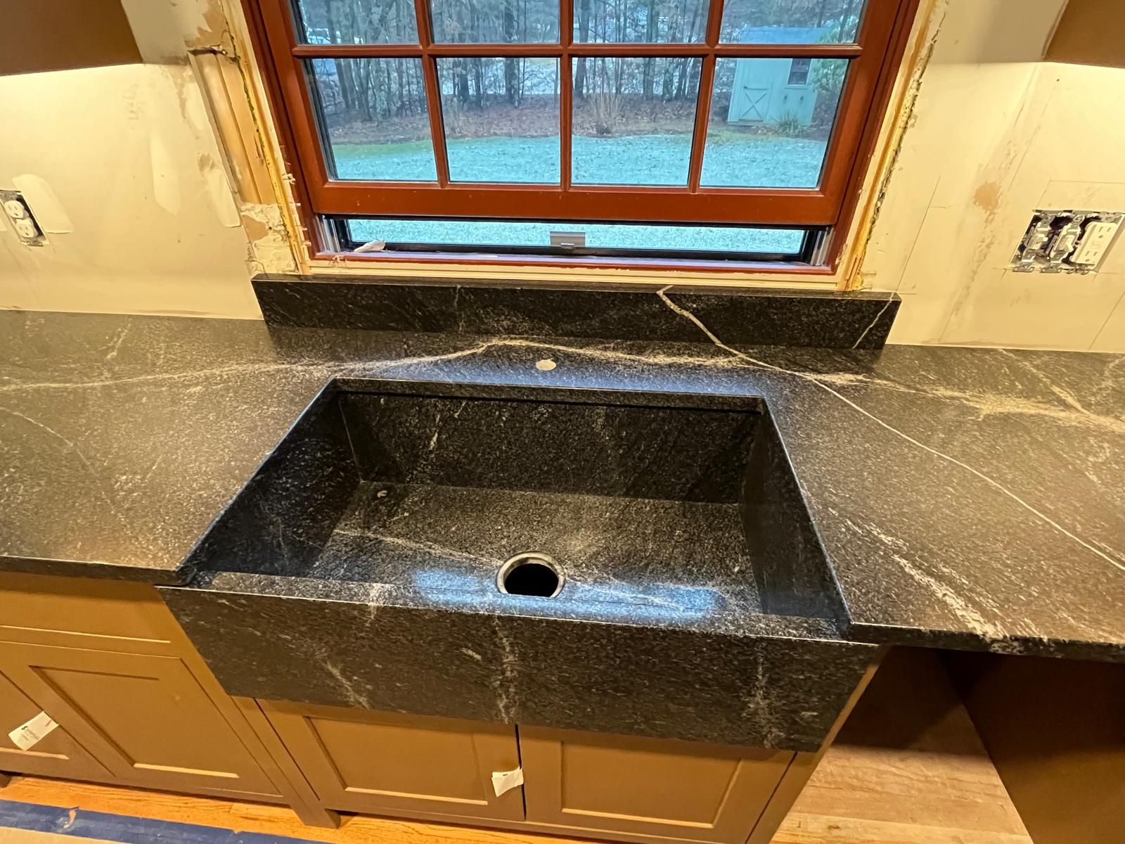 Black stone sink and countertop installed under a window in a kitchen, with tan cabinets below.