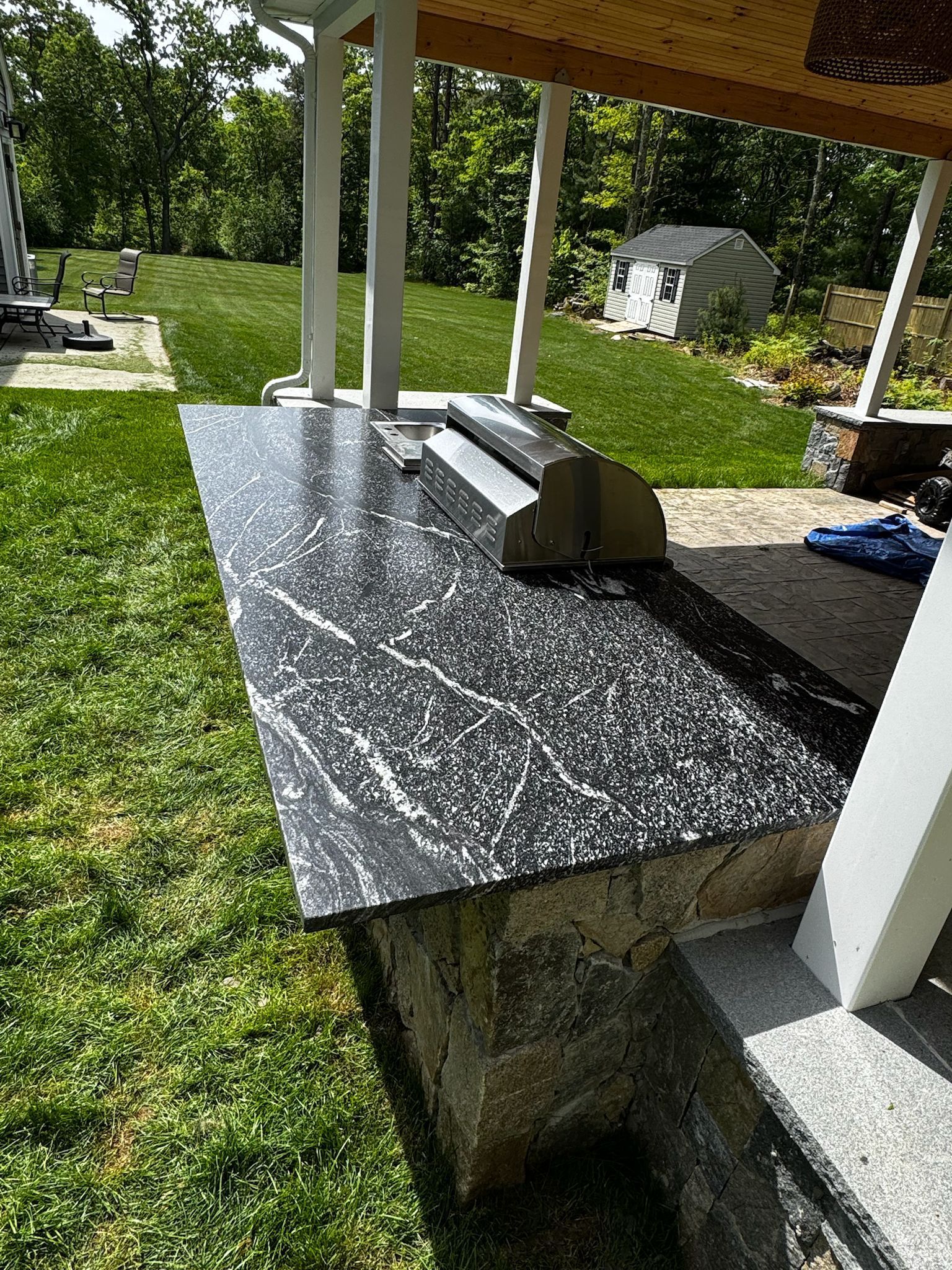 Outdoor kitchen with black and white speckled countertop and built-in grill, under a covered patio.