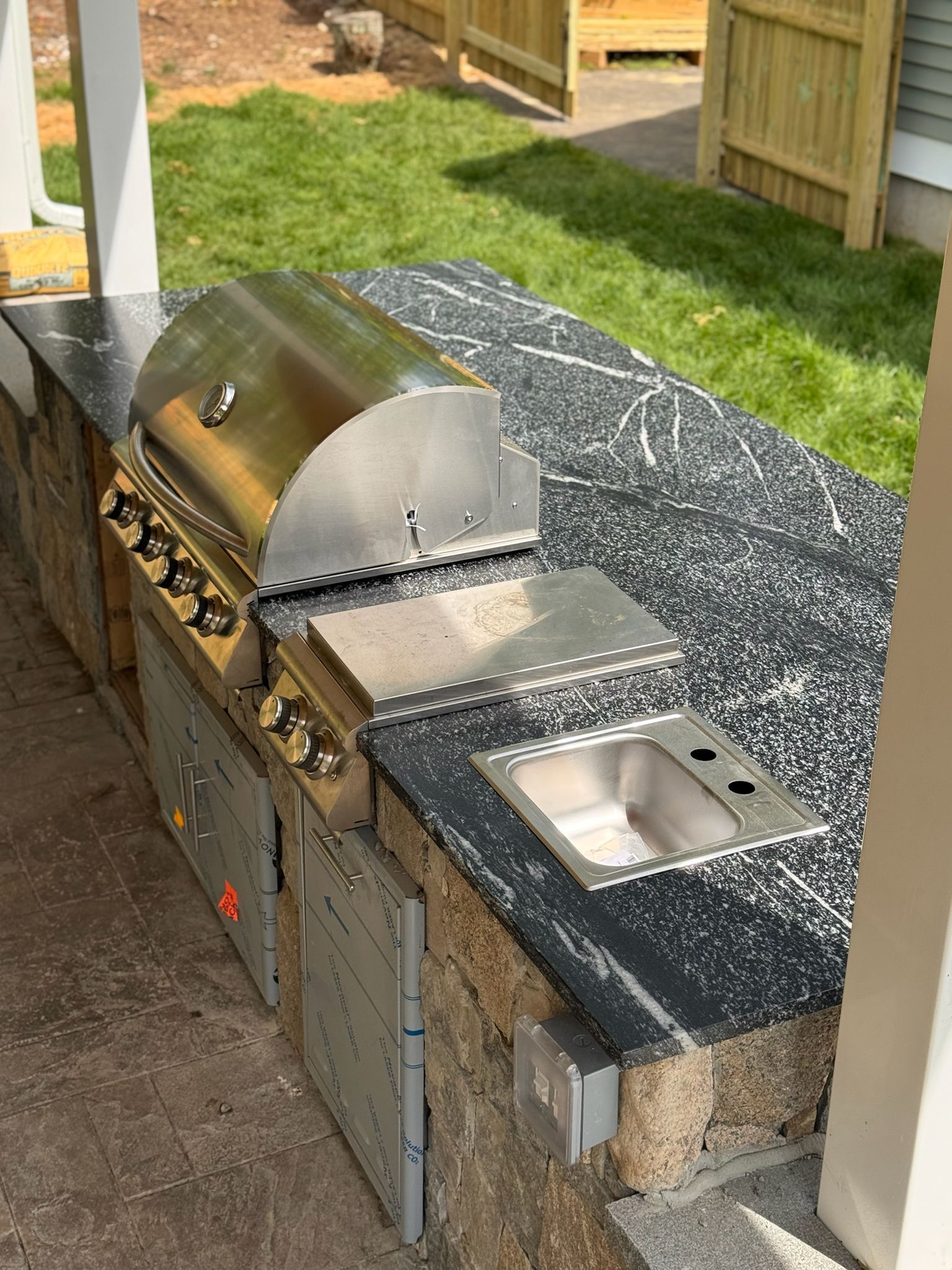 Outdoor kitchen with a stainless steel grill, sink, and cabinets on a speckled black countertop.