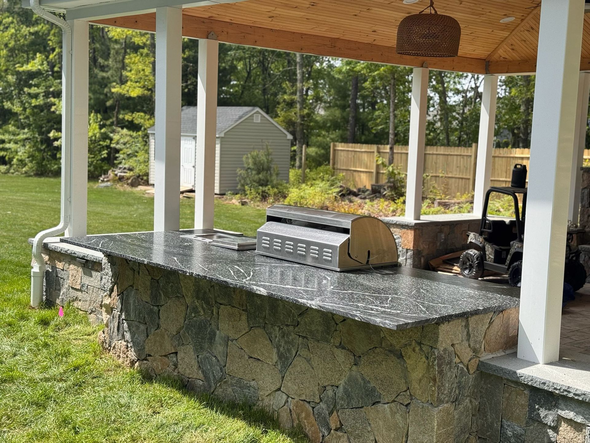 Outdoor kitchen with stone base, granite countertop, grill, under covered patio. Green grass, shed in background.