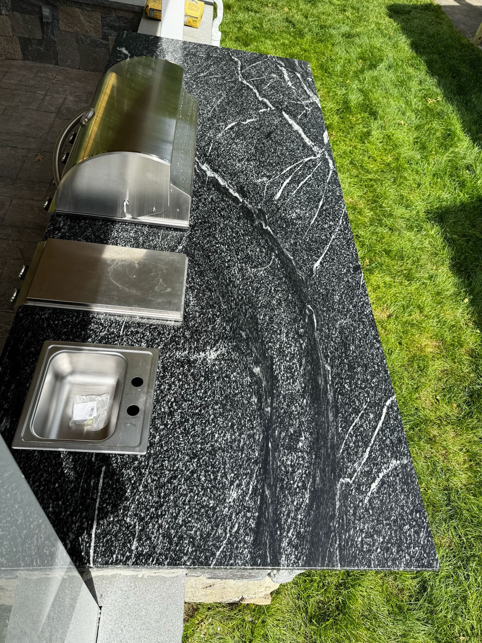 Outdoor kitchen countertop with stainless steel grill, sink, and black speckled surface.