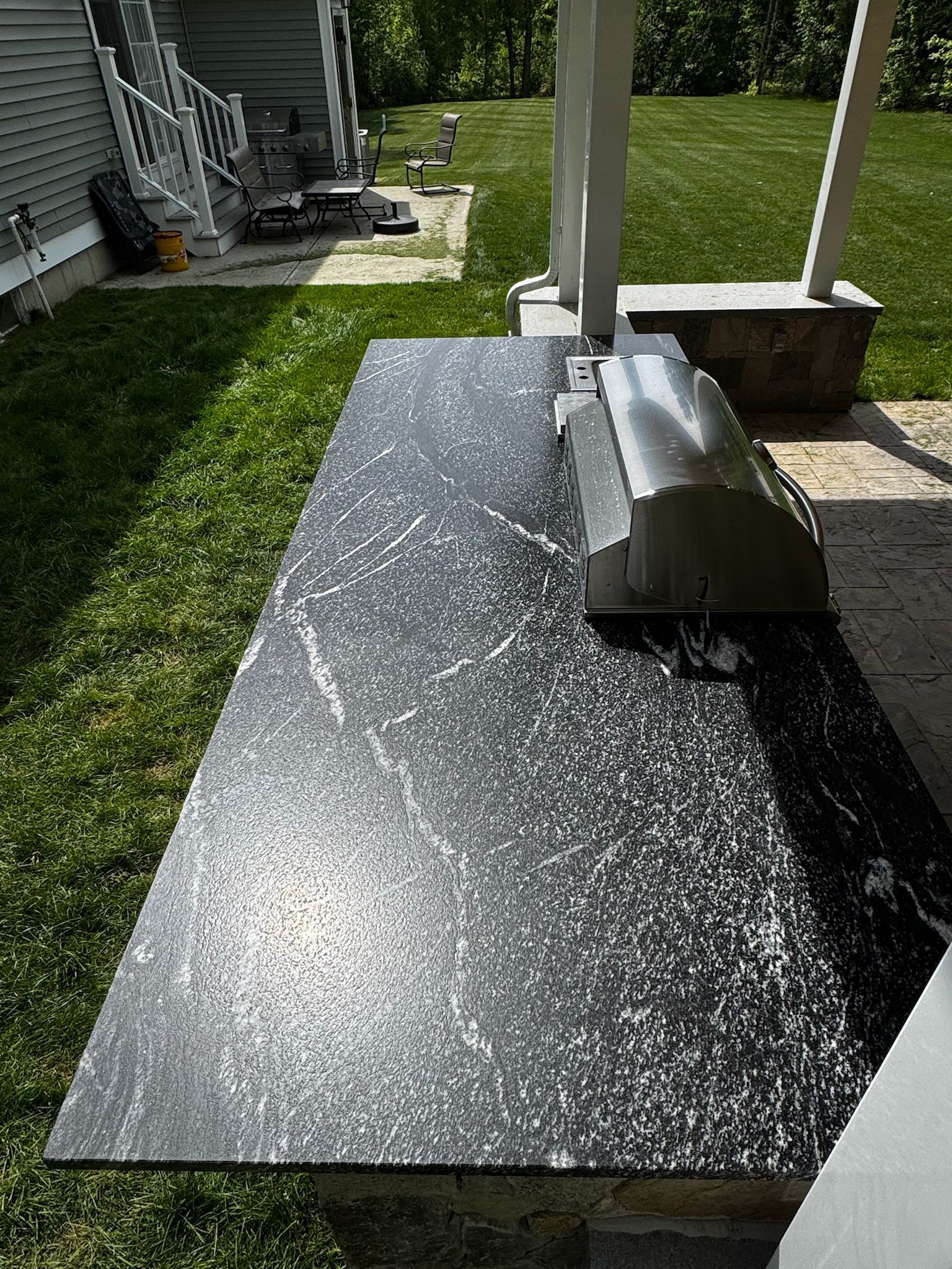 Outdoor kitchen countertop with built-in grill, granite-like finish. Green grass and house in background.