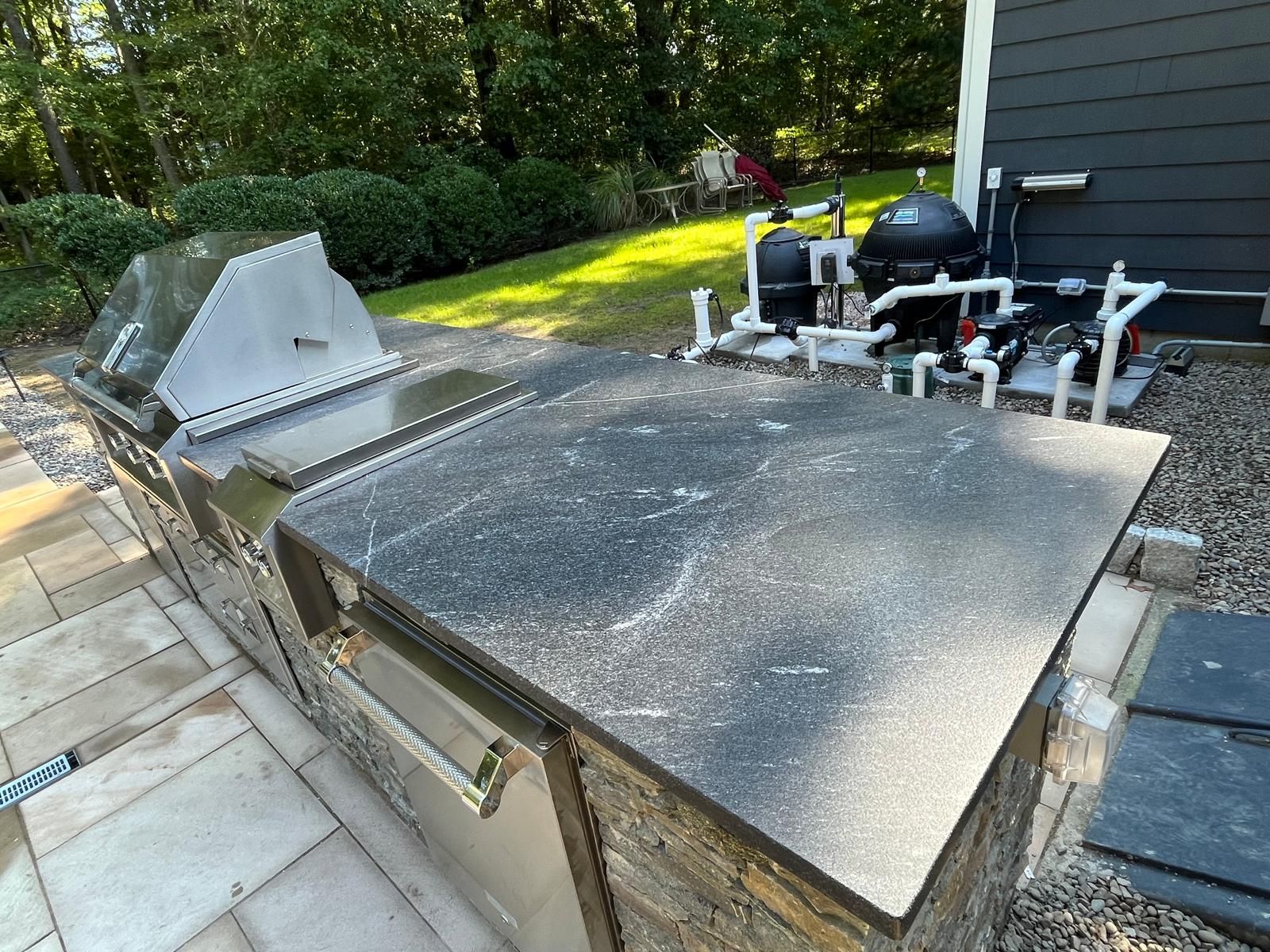 Outdoor kitchen with stainless steel grill, dark countertop, and stone facade, adjacent to a pool equipment area.