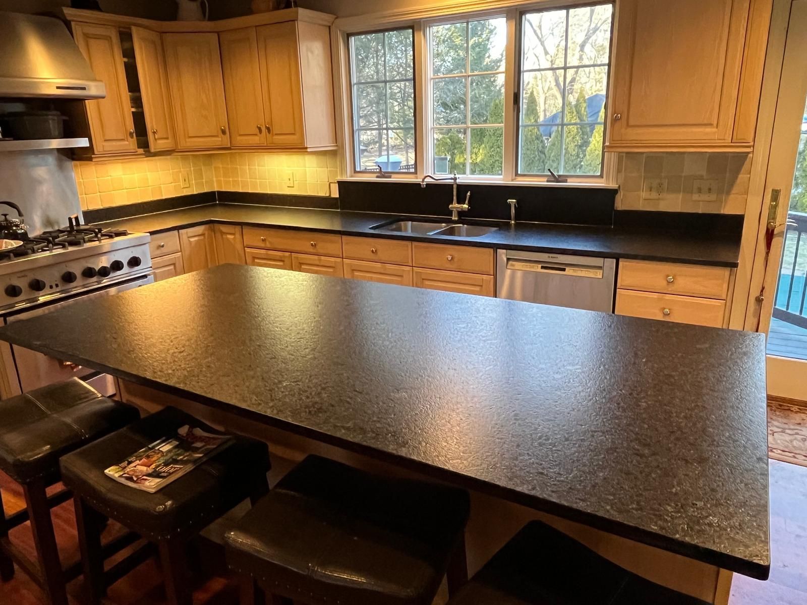 Kitchen with black countertops, light wood cabinets, stainless steel appliances, and a large island with stools.
