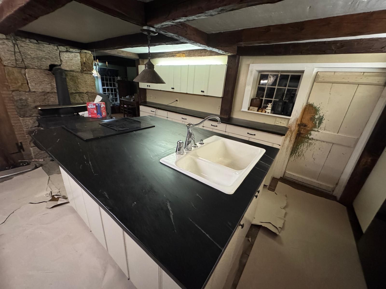 Kitchen with black countertop, white cabinets, sink, and window. Wooden beams and door in a rustic setting.