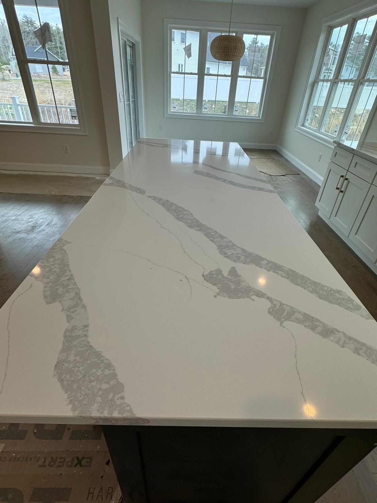 Large kitchen island with white countertop, grey veining, and dark base. Windows in the background.
