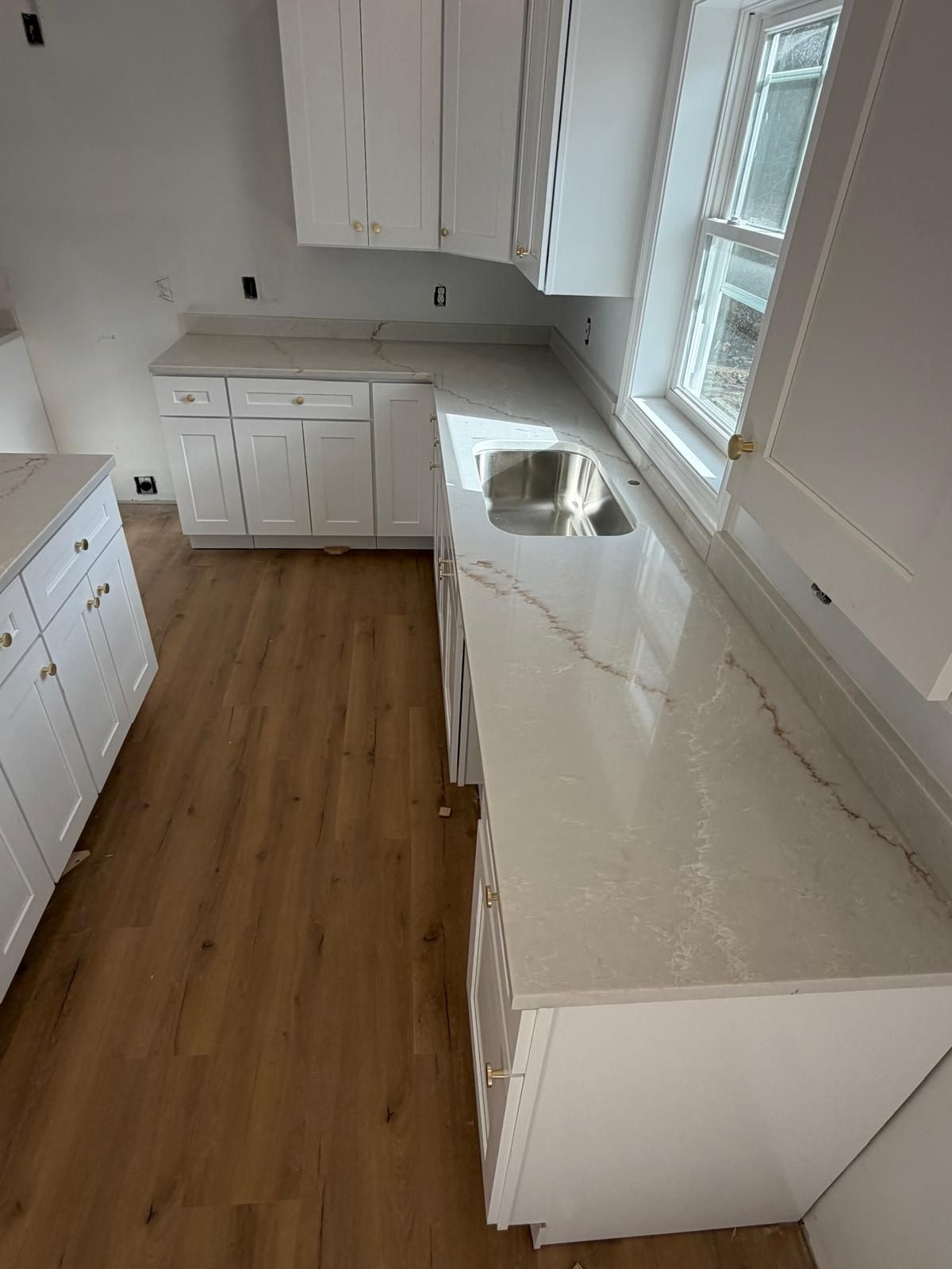 White kitchen cabinets and countertops, wood flooring. Sink next to a window.