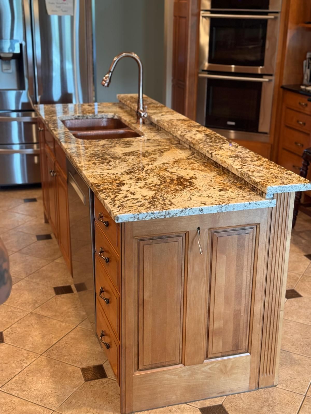 Kitchen island with granite countertop, sink, faucet, and wooden cabinetry. Stainless steel appliances in background.