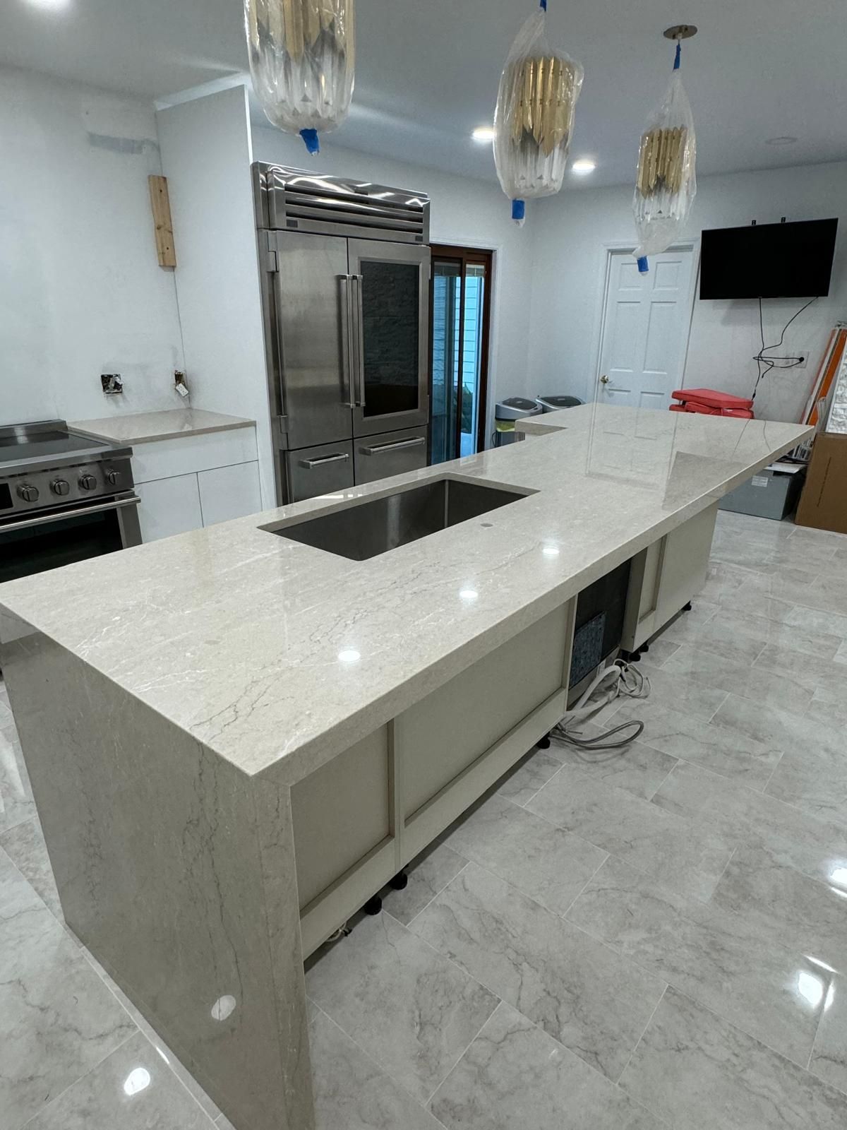 Modern kitchen with large island, stainless steel refrigerator, and pendant lights.