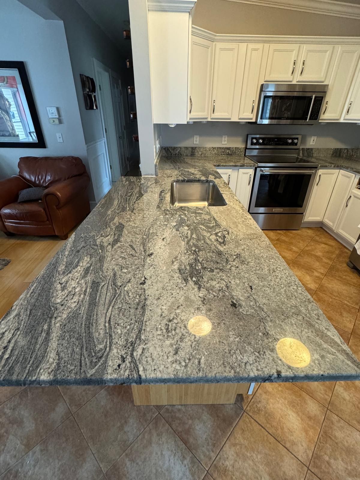 Kitchen island with granite countertop, sink, and white cabinets. Stainless steel appliances and leather armchair.