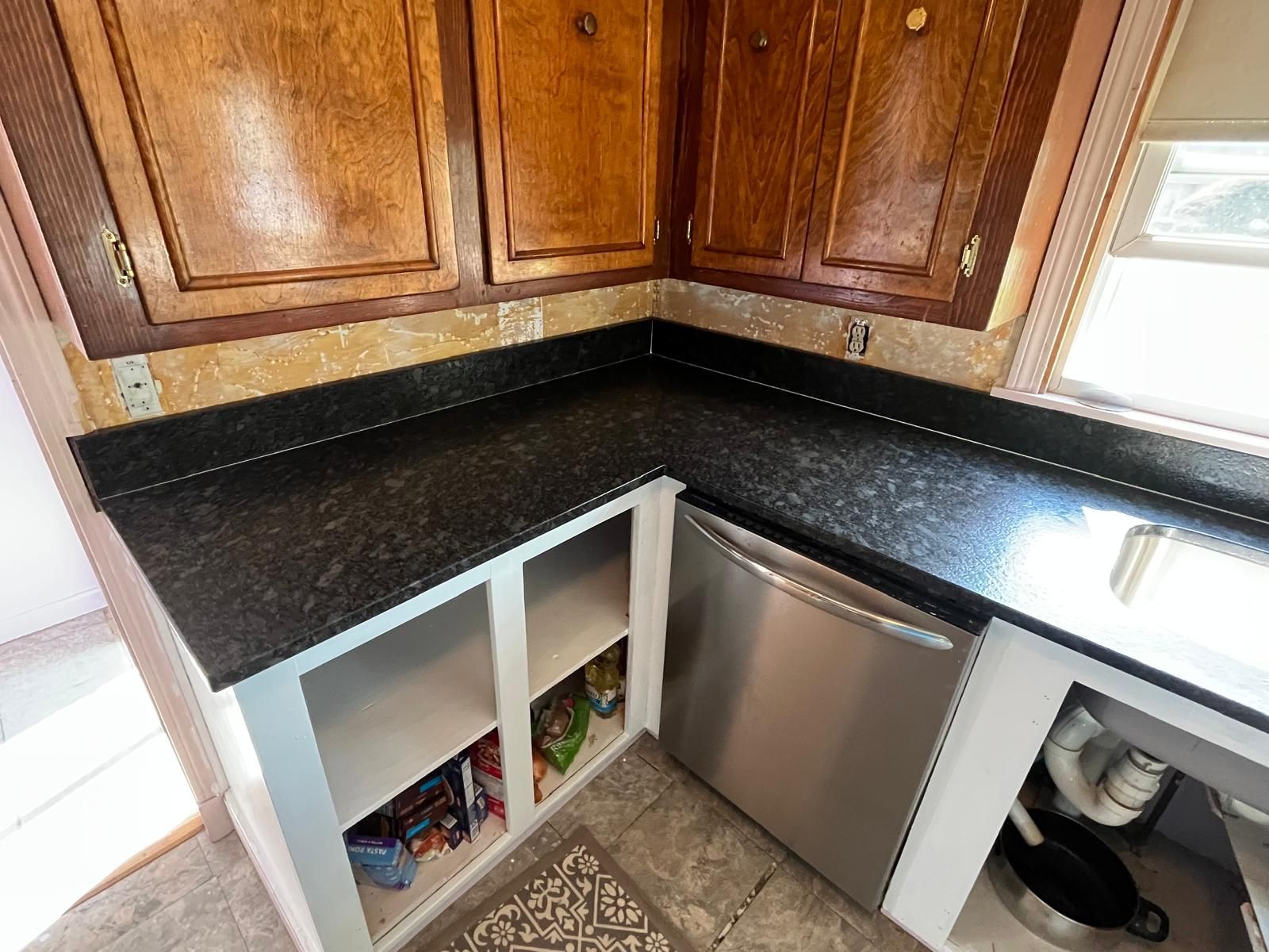 Kitchen corner with dark countertops, stainless steel dishwasher, and wooden cabinets.