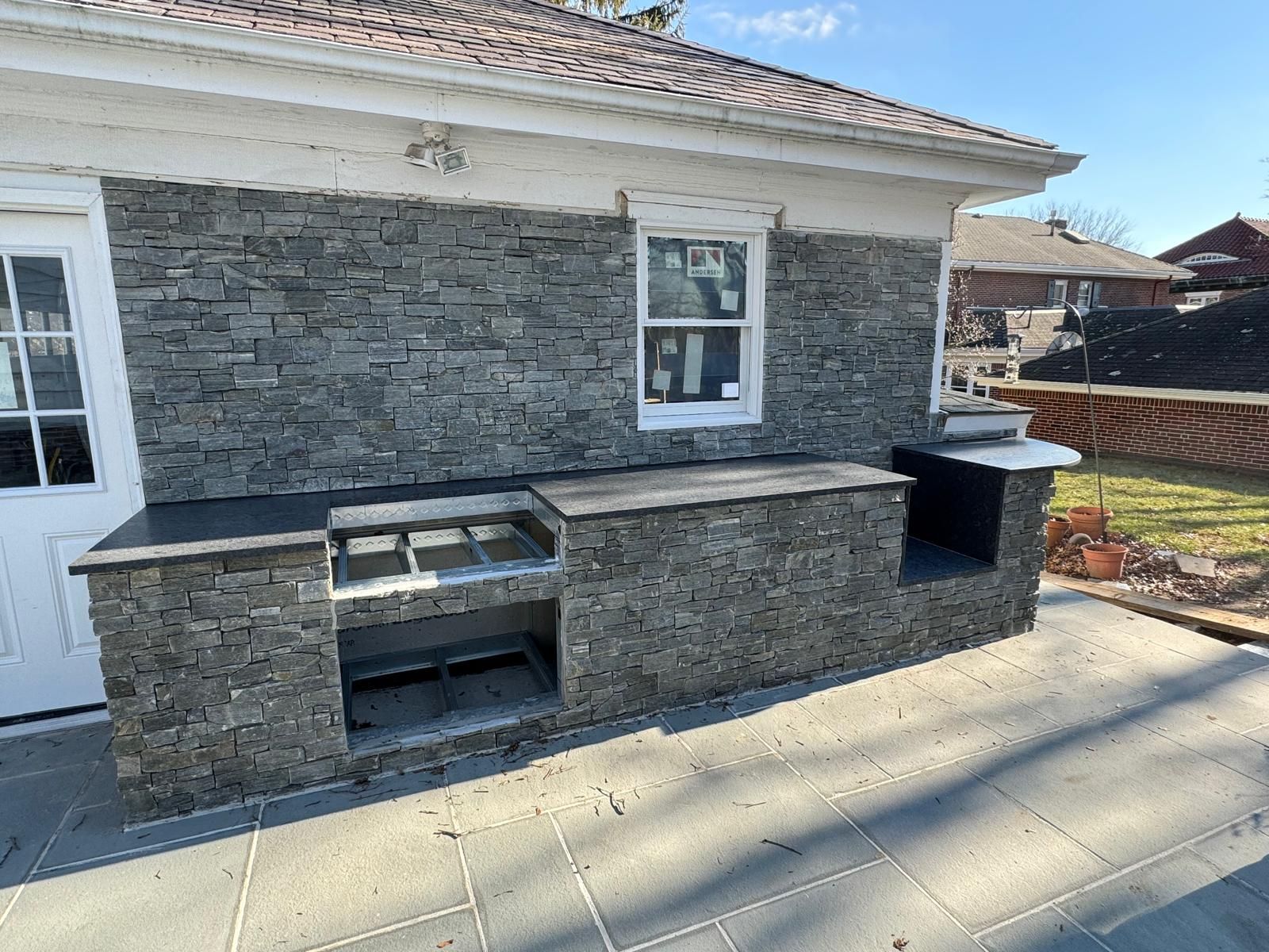 Outdoor kitchen with gray stone veneer and dark countertops. Includes a built-in grill and a small window.