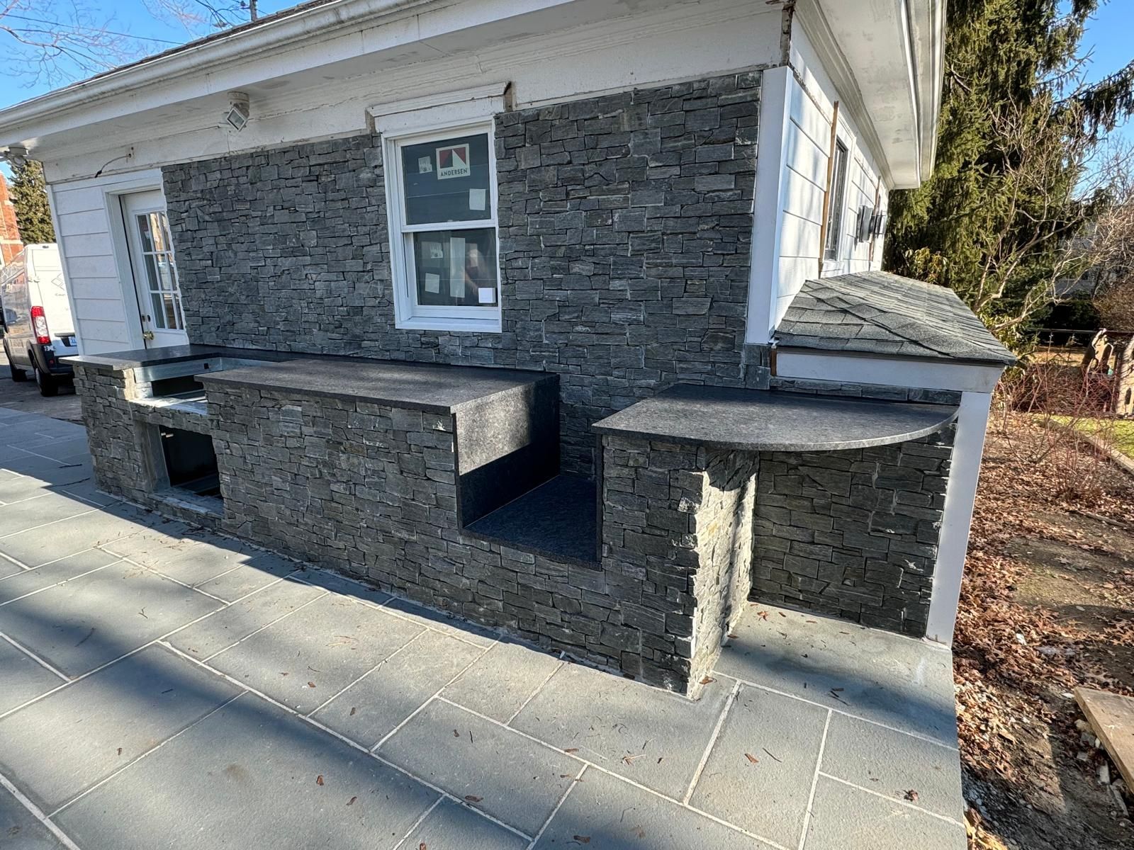 Outdoor kitchen with dark stone facade and countertops; built-in grill and prep areas; gray paving.
