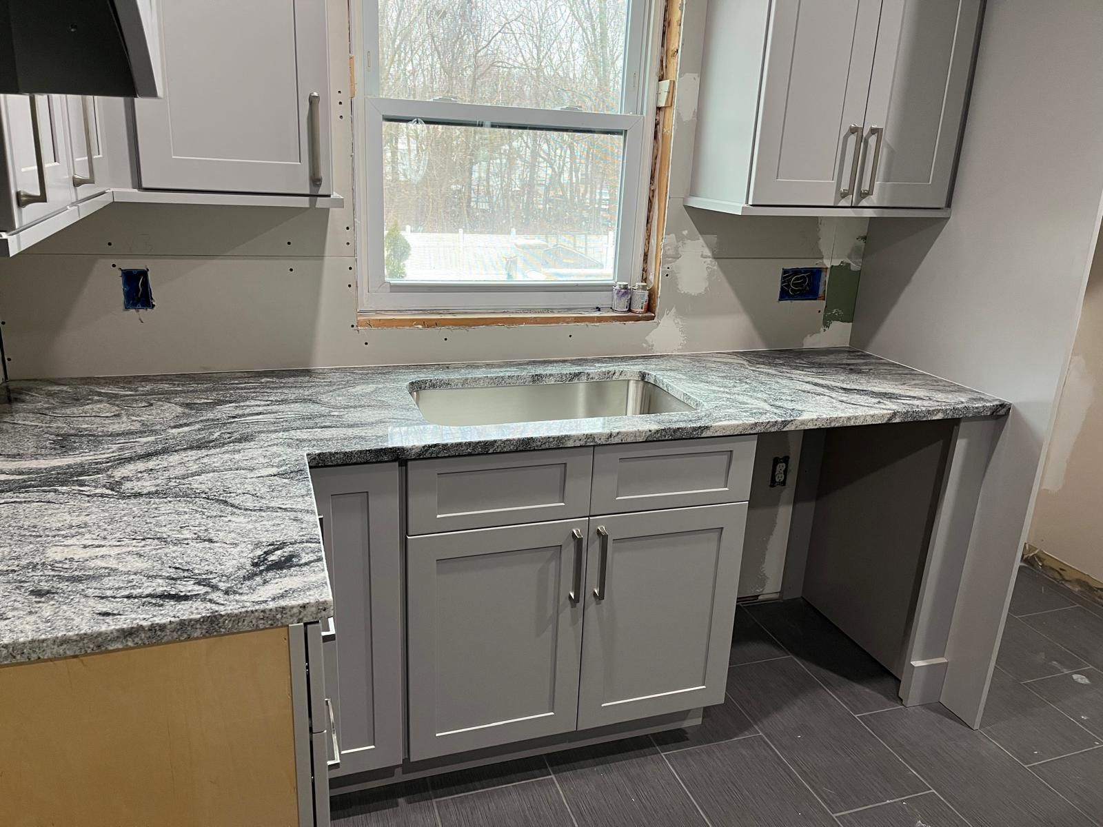Gray kitchen cabinets with granite countertop, sink under window, and unfinished walls.