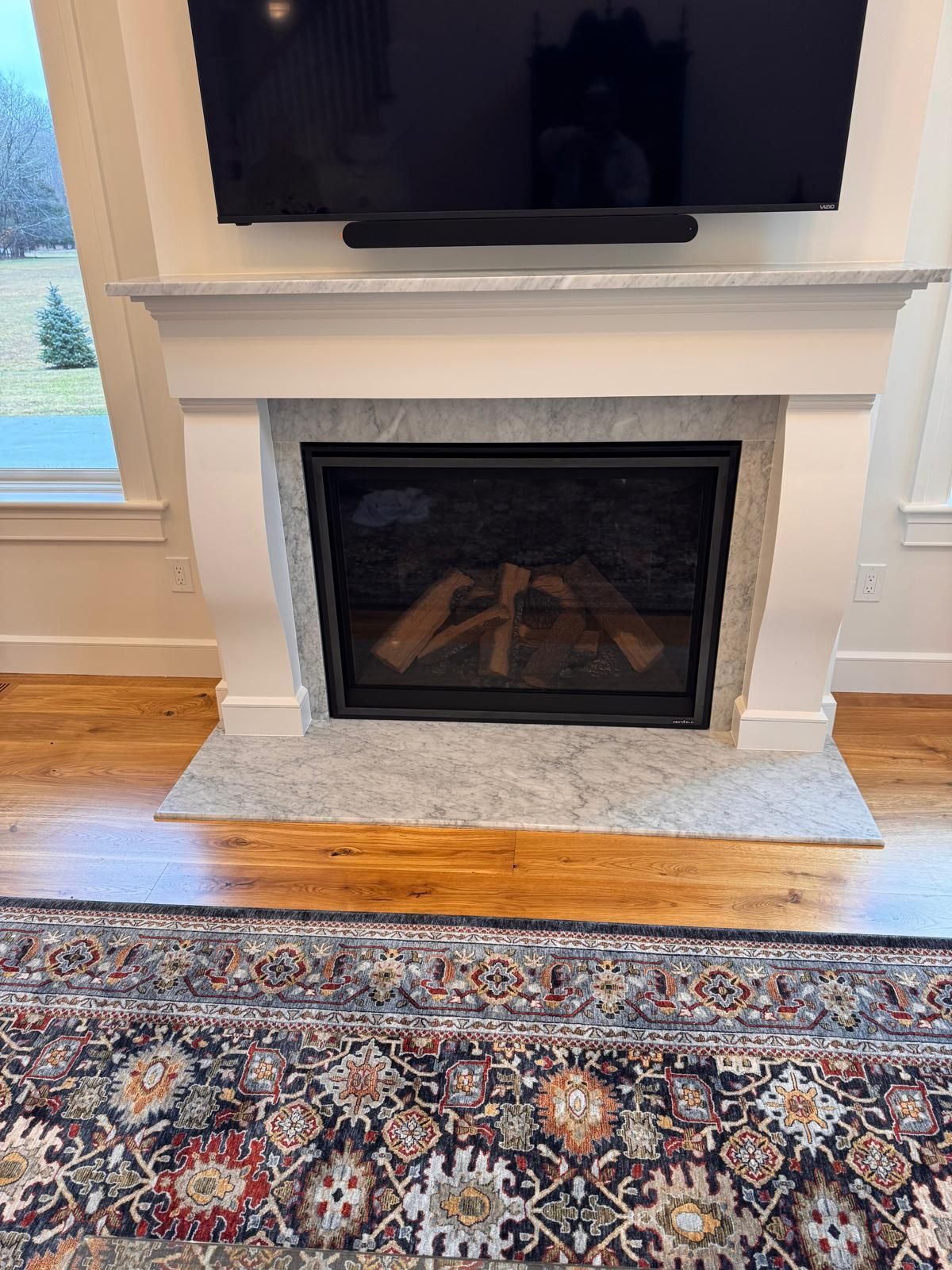 Fireplace with wood logs, marble, a large TV above, and ornate rug on hardwood floor.