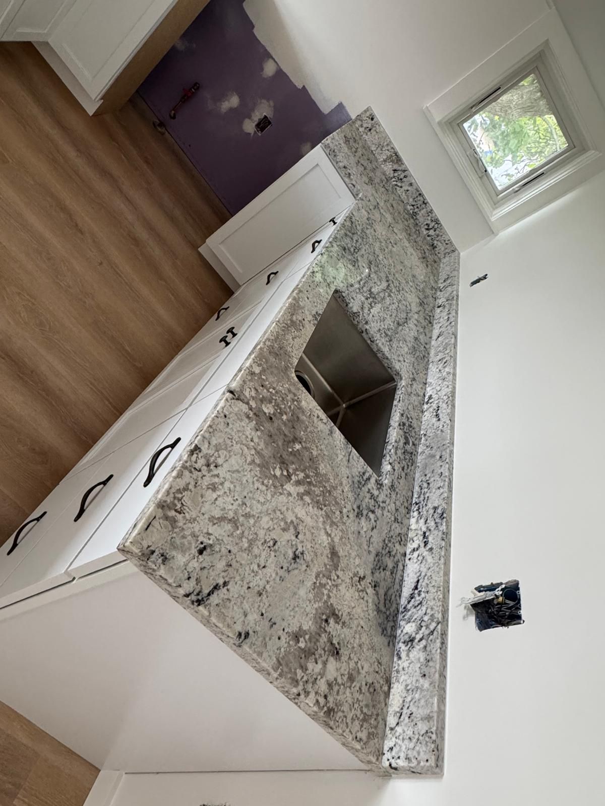 Kitchen island with granite countertop and sink. White cabinets, light wood floor, small window.
