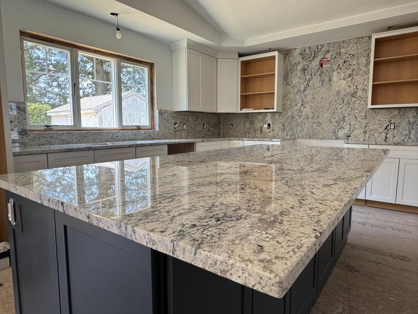 Kitchen with a large granite island and countertops. White and grey granite with dark cabinets and unfinished white cabinets.