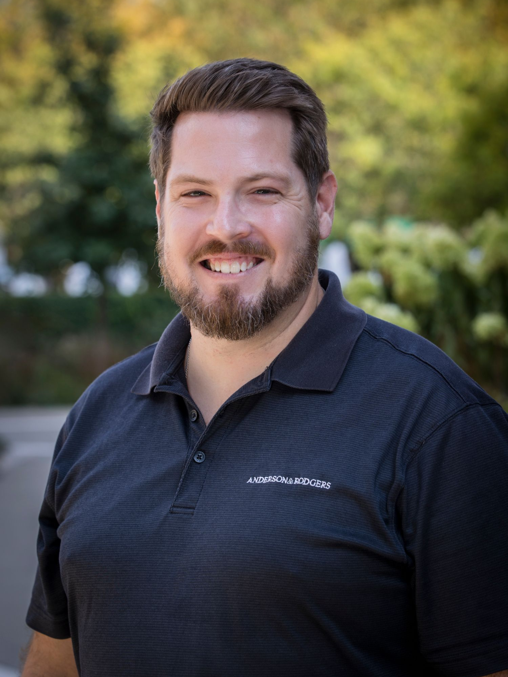 A man with a beard is wearing a blue vest and smiling for the camera.