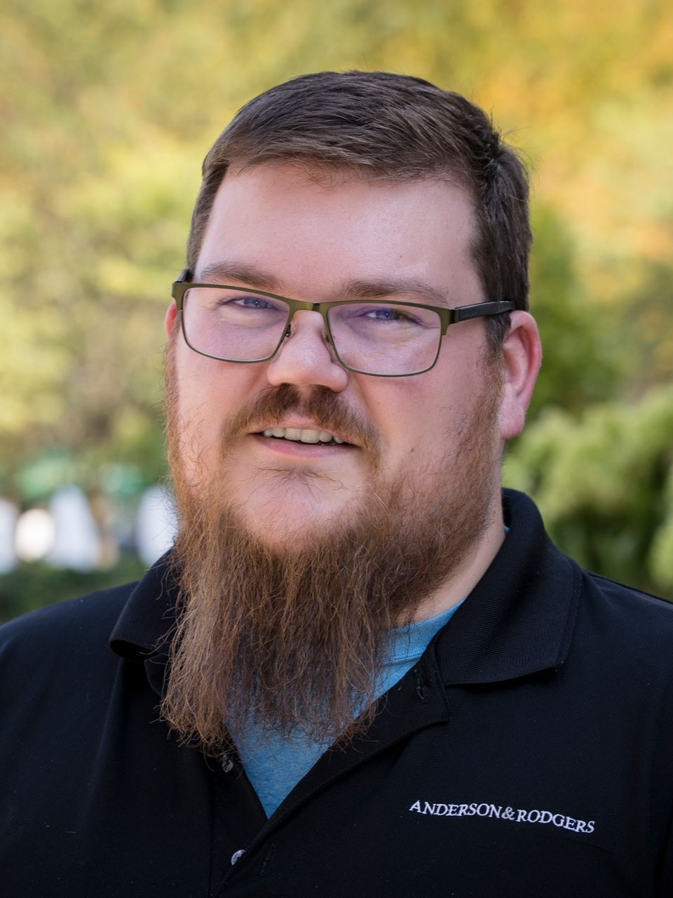 A man with a beard wearing glasses and a blue shirt is standing in front of a building.