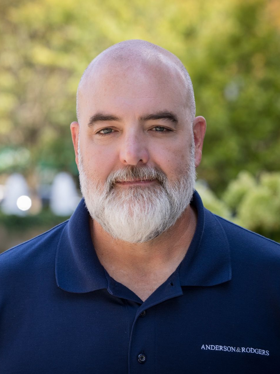 A bald man with a beard is wearing a plaid shirt and a black vest.