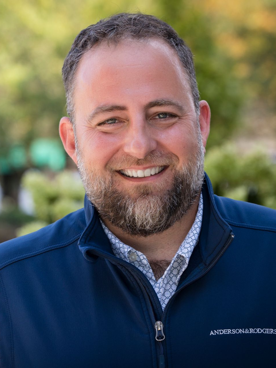 A man with a beard is wearing a blue vest and smiling for the camera.