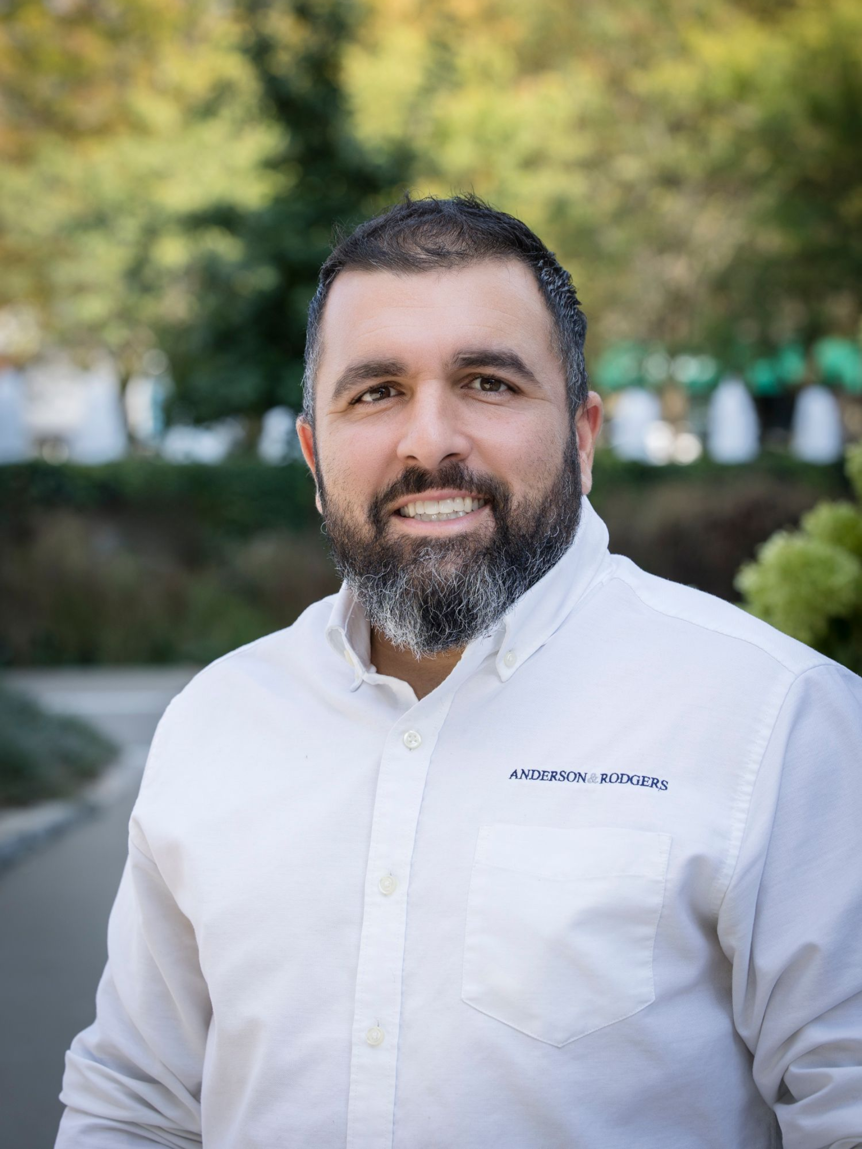 A man with a beard is wearing a blue and white checkered shirt and smiling.
