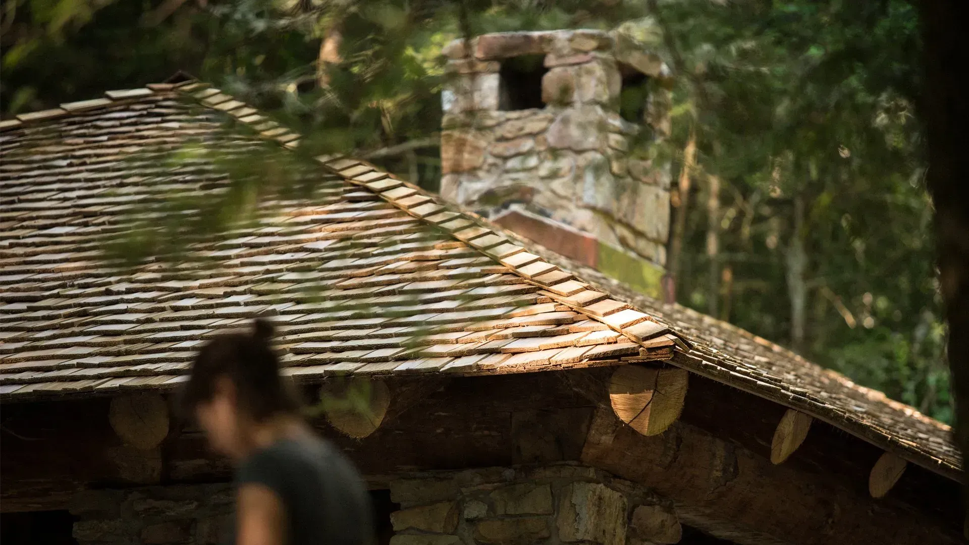 A woman is standing in front of a wooden house with a chimney on the roof.