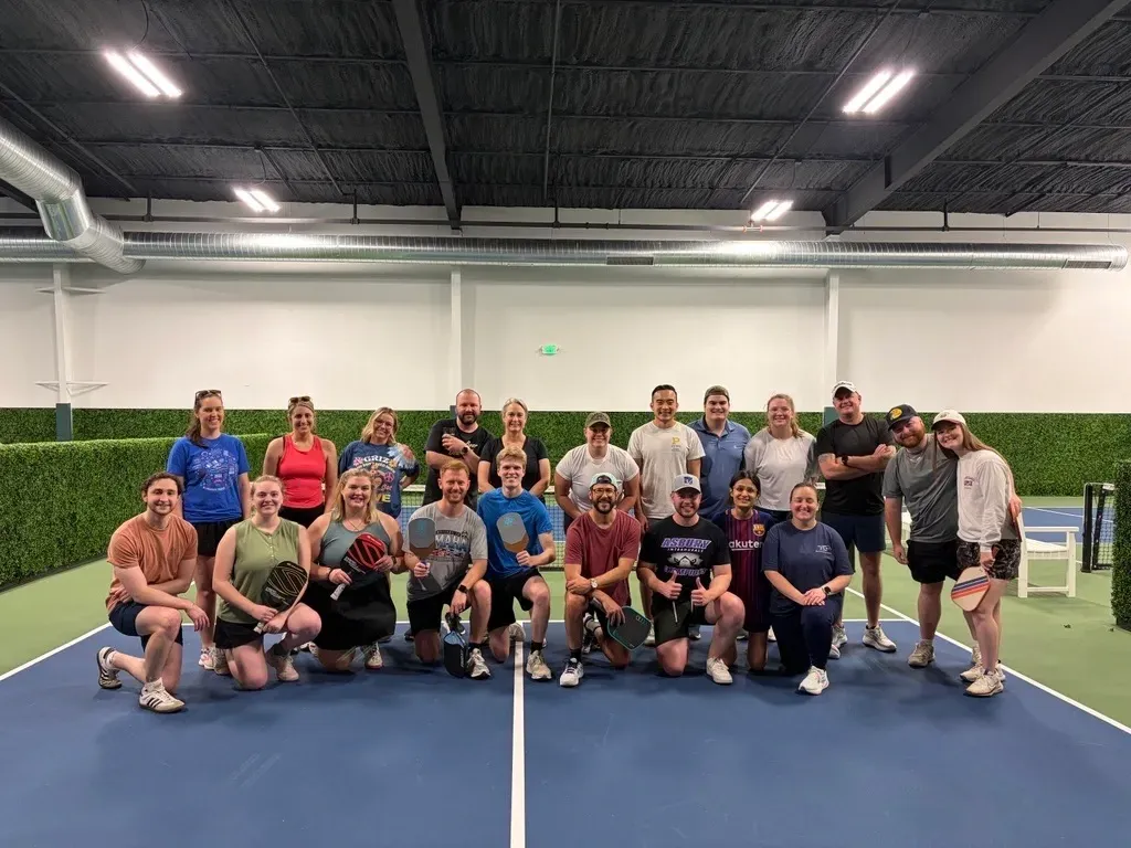 Group of people posing on an indoor tennis court under bright lights, with hedge walls in the background