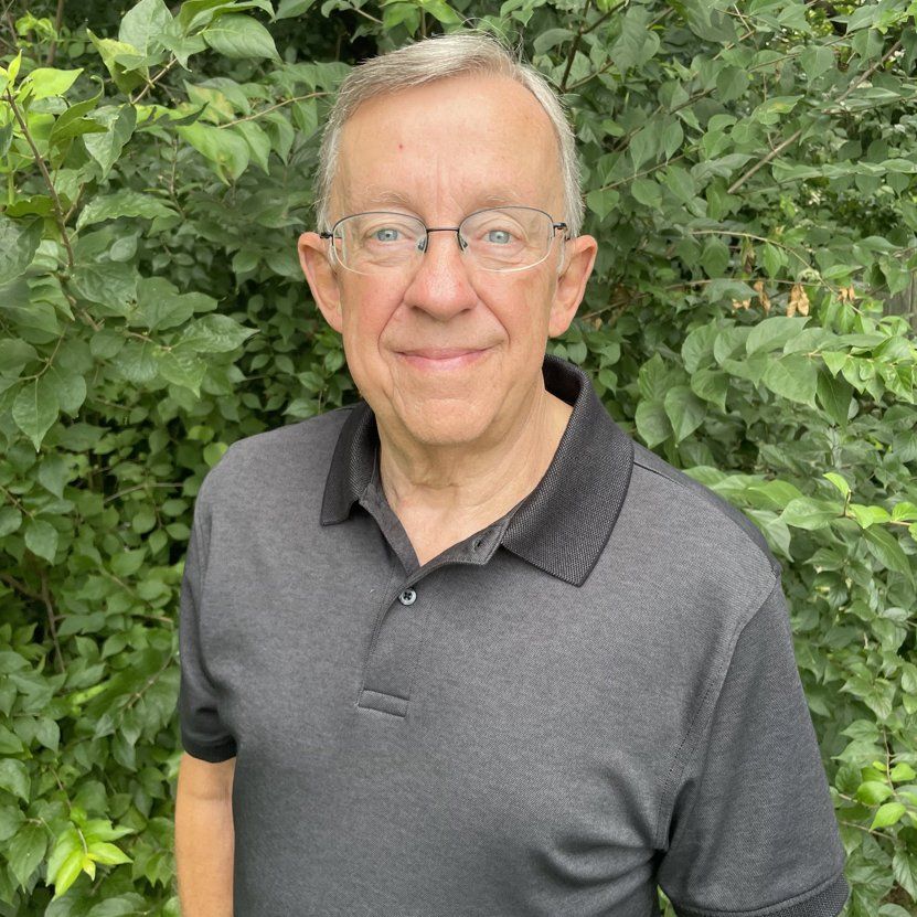 A man wearing glasses and a black shirt is standing in front of a bush.