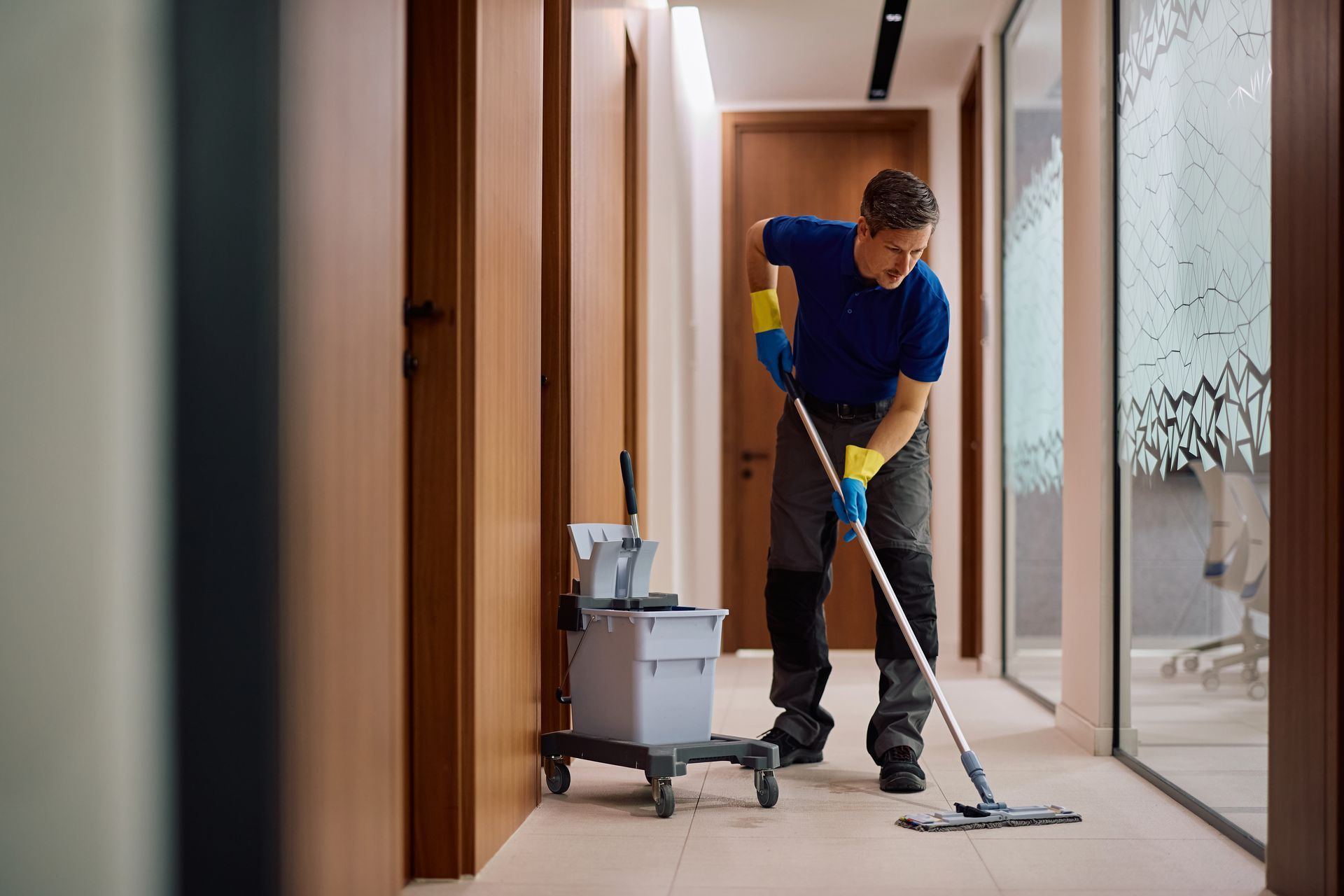 Cleaner mopping a hallway floor beside a cleaning cart in a modern office corridor.