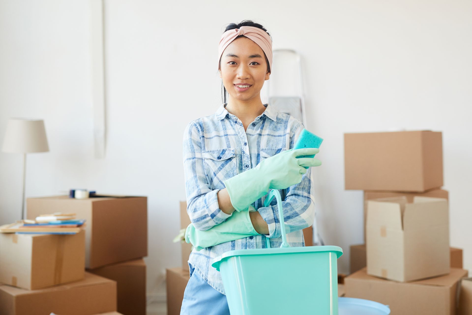 Person in gloves cleaning a home with spray bottle and bucket, surrounded by moving boxes.