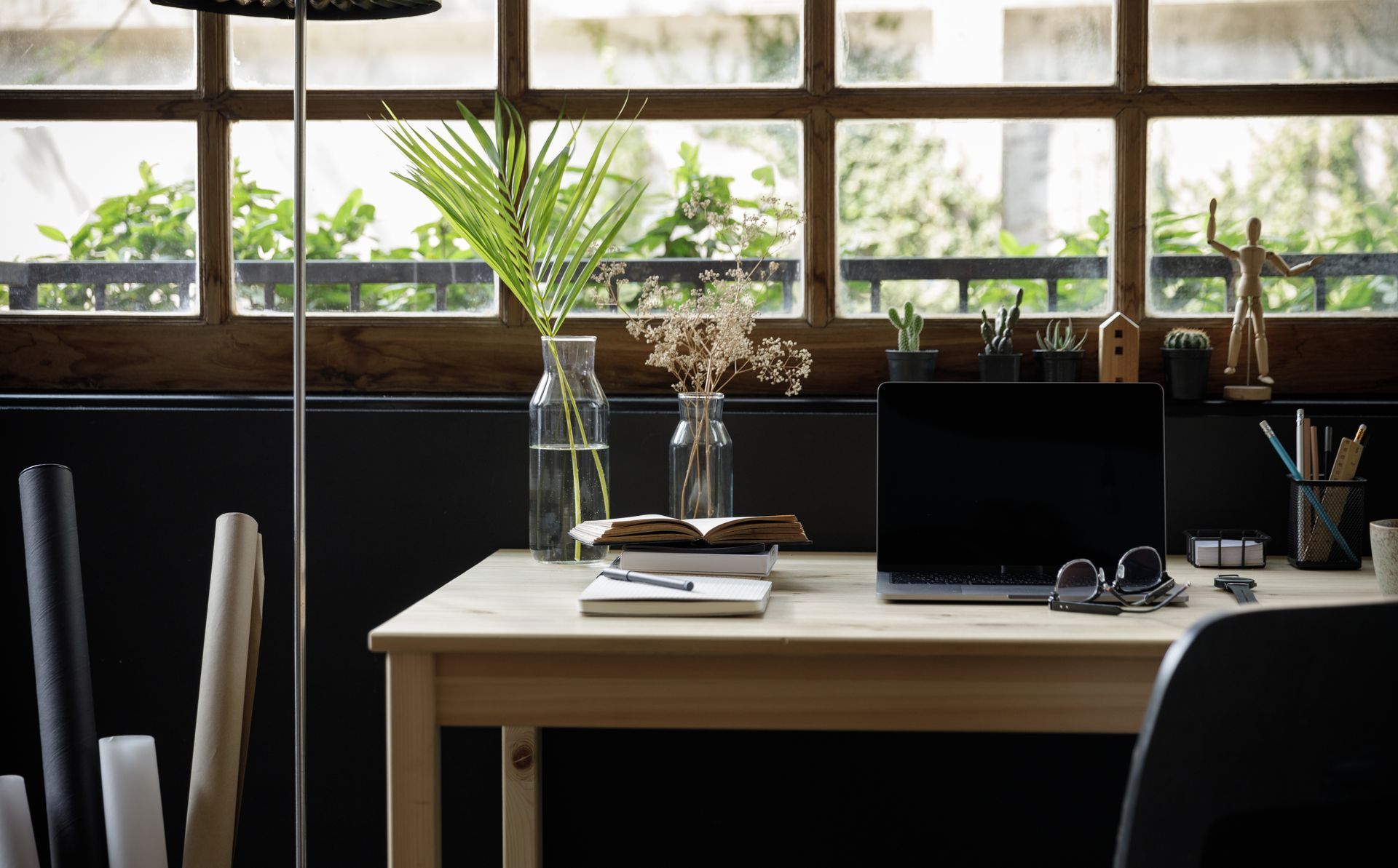 Sunlit office desk by large windows with plants, laptop, and office chair