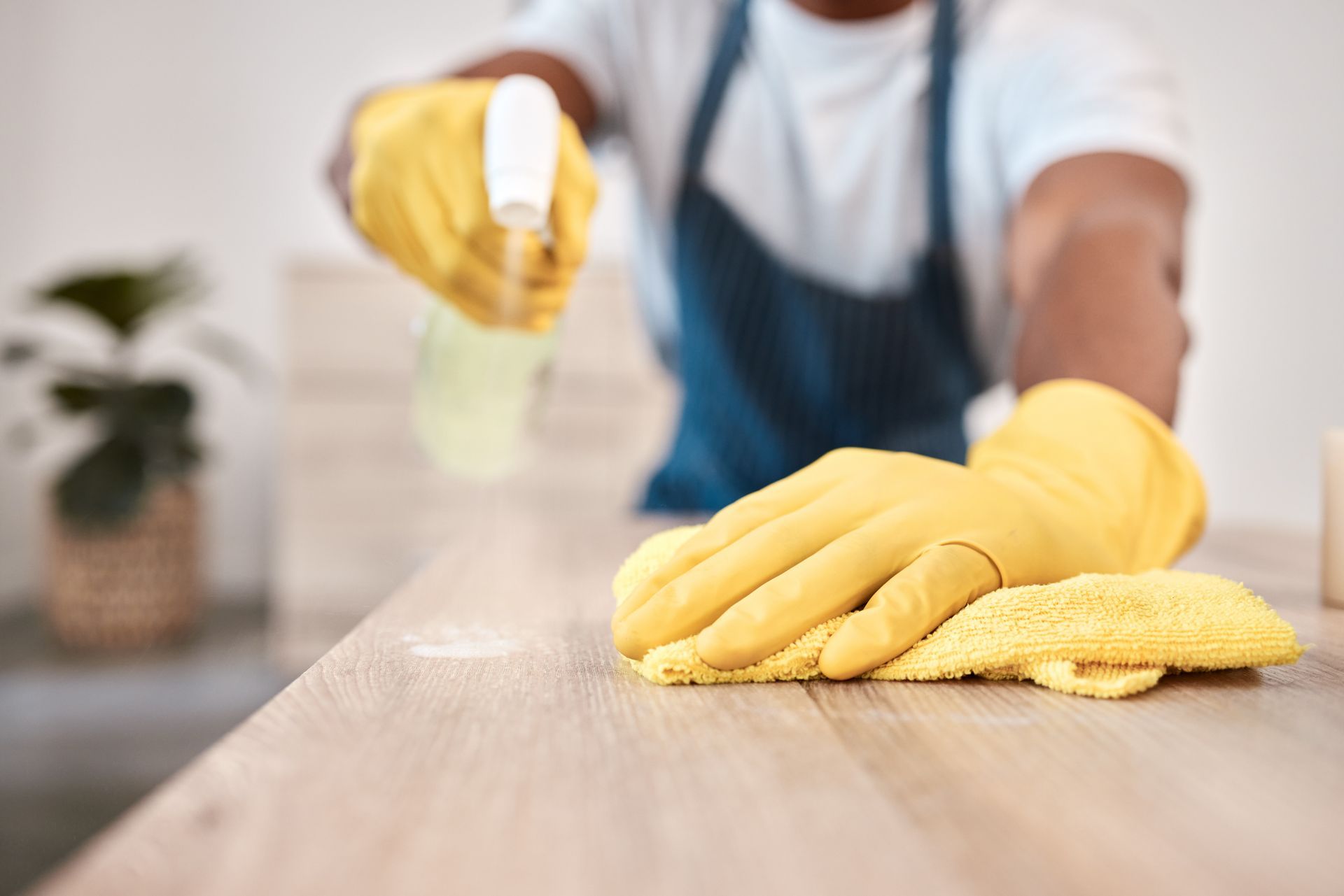 Person in gloves wiping a wooden table with a yellow cloth in a kitchen