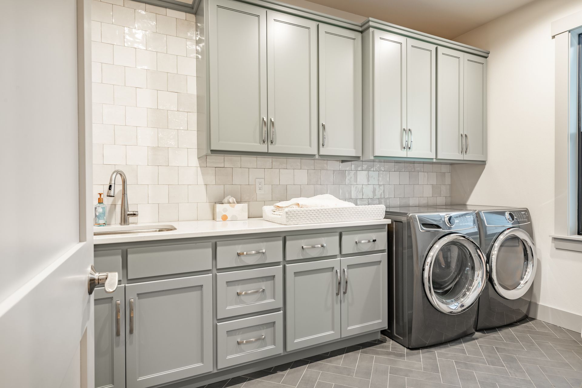 Laundry room with gray cabinets, white backsplash, and gray washer/dryer.