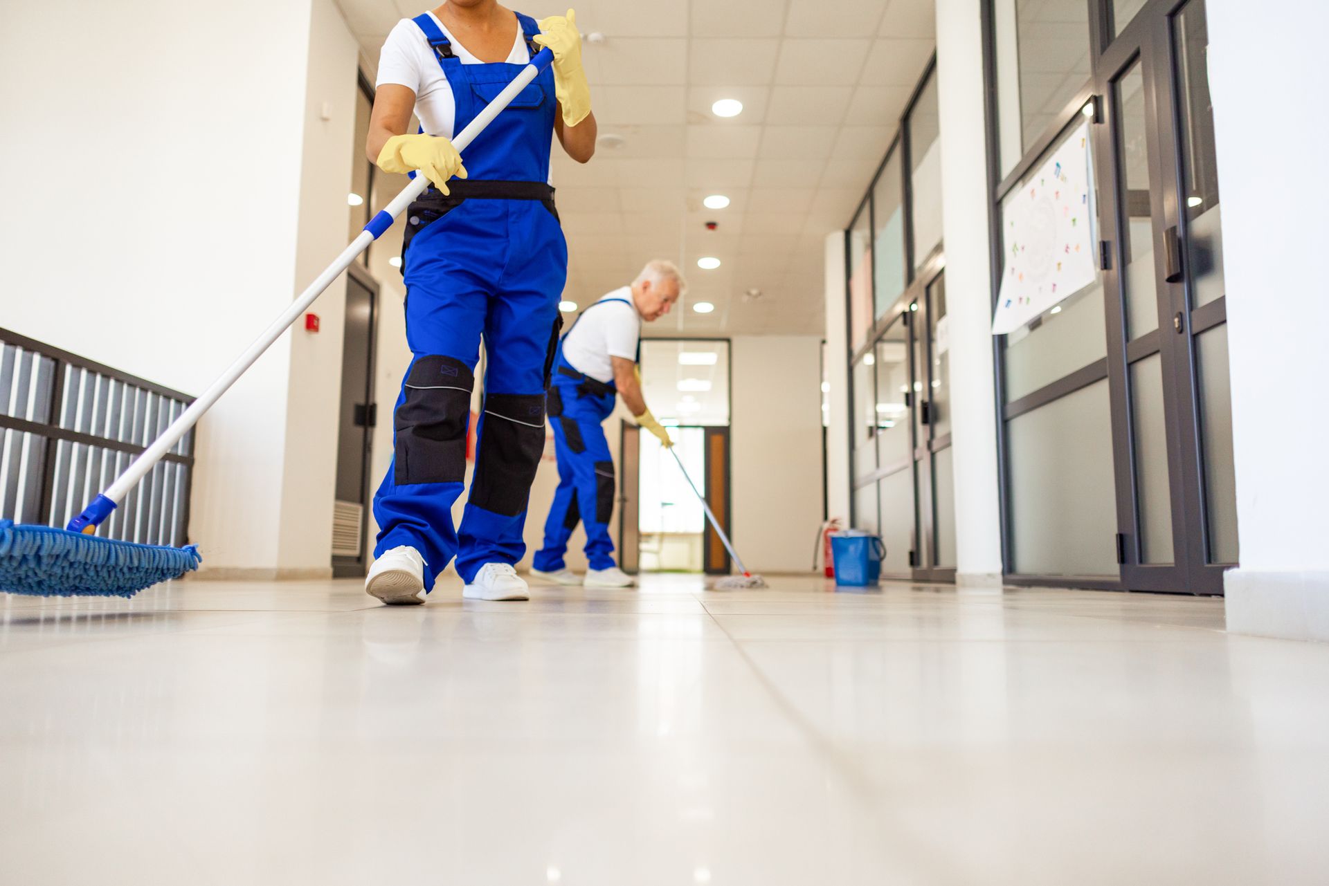Two people mopping a clean hallway. They wear blue coveralls and yellow gloves.