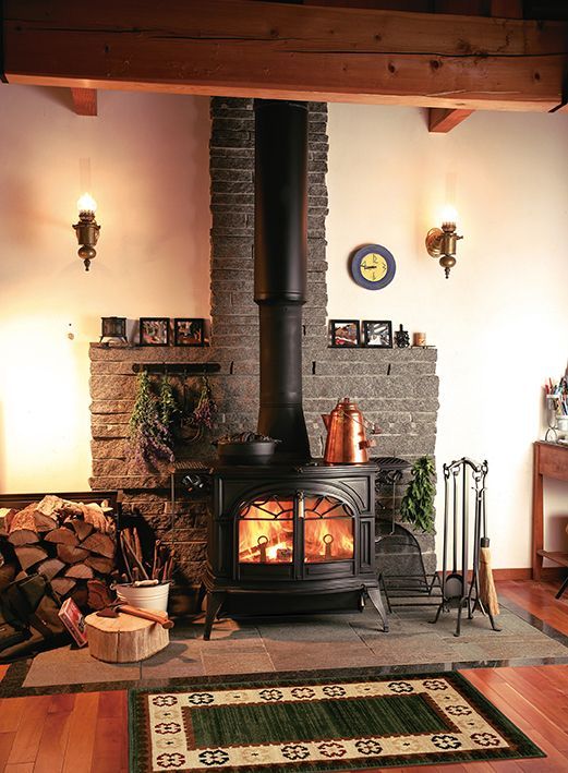 Cozy living room with a lit wood stove, stacked firewood, and a patterned rug beneath exposed wooden beams