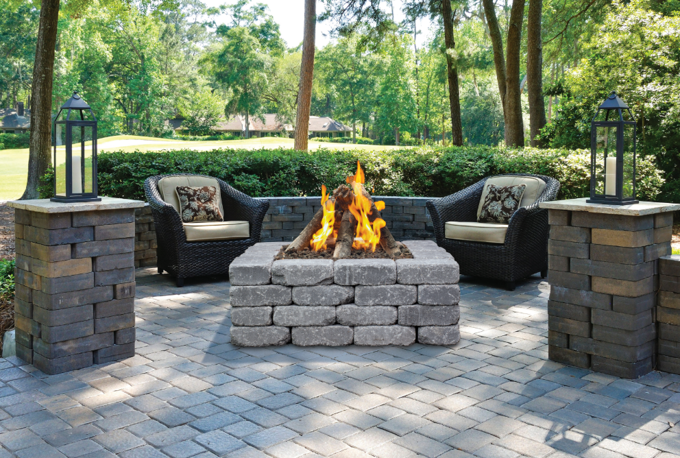 Custom stone outdoor fire pit with flames, surrounded by cushioned chairs on a patio with greenery beyond