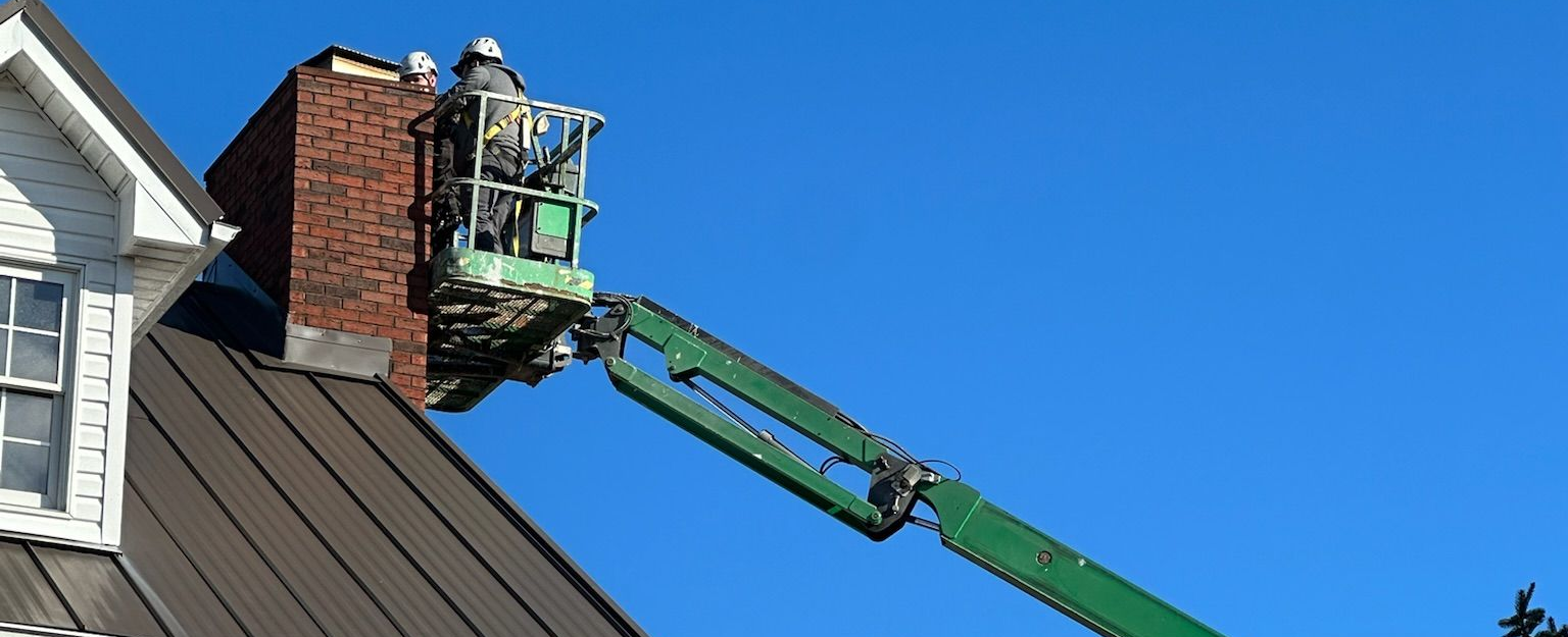 Worker in a green boom lift repairing a brick chimney on a sloped roof under a blue sky