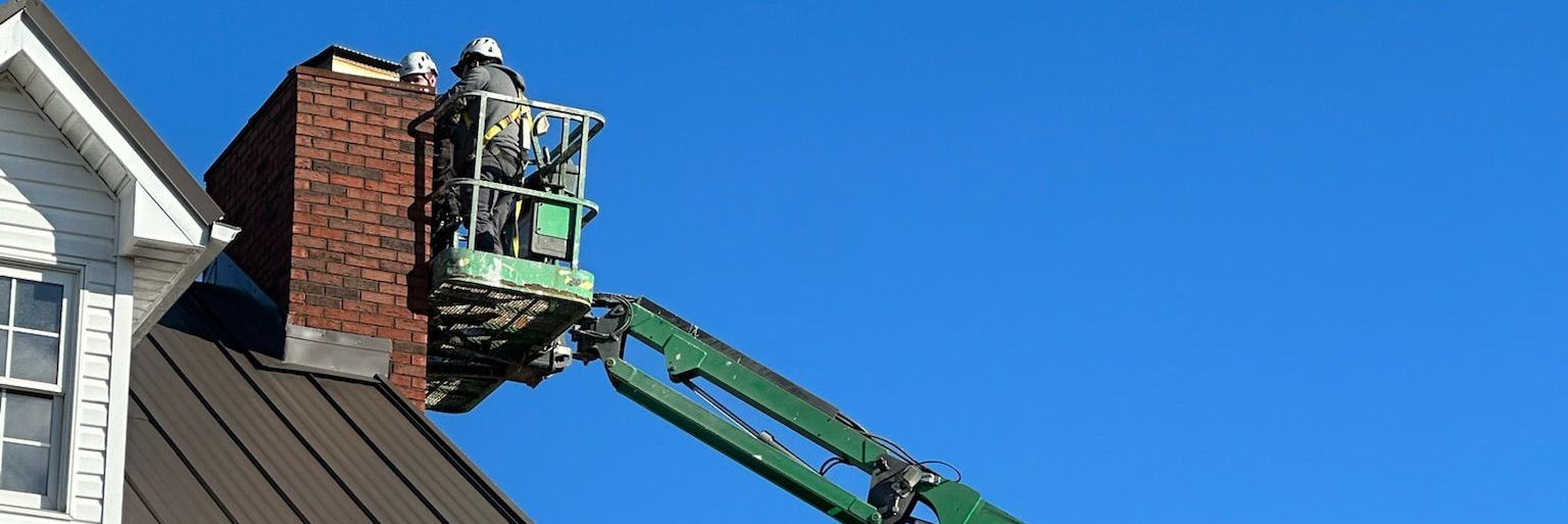 Green boom lift reaches a red brick chimney on a rooftop under a clear blue sky