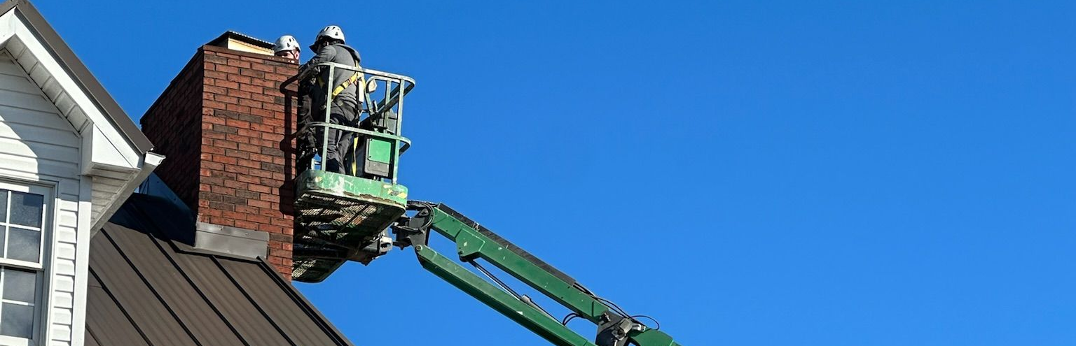 Worker in a green boom lift repairing a brick chimney on a sloped roof under a blue sky