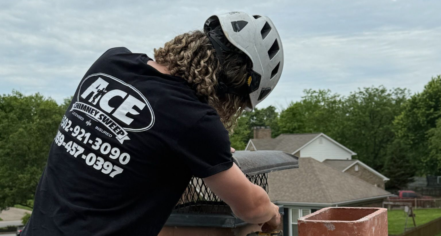 Chimney technician in helmet inspects a roof, wearing a black shirt with Ace Chimney Fireplace & Outdoor Living logo.