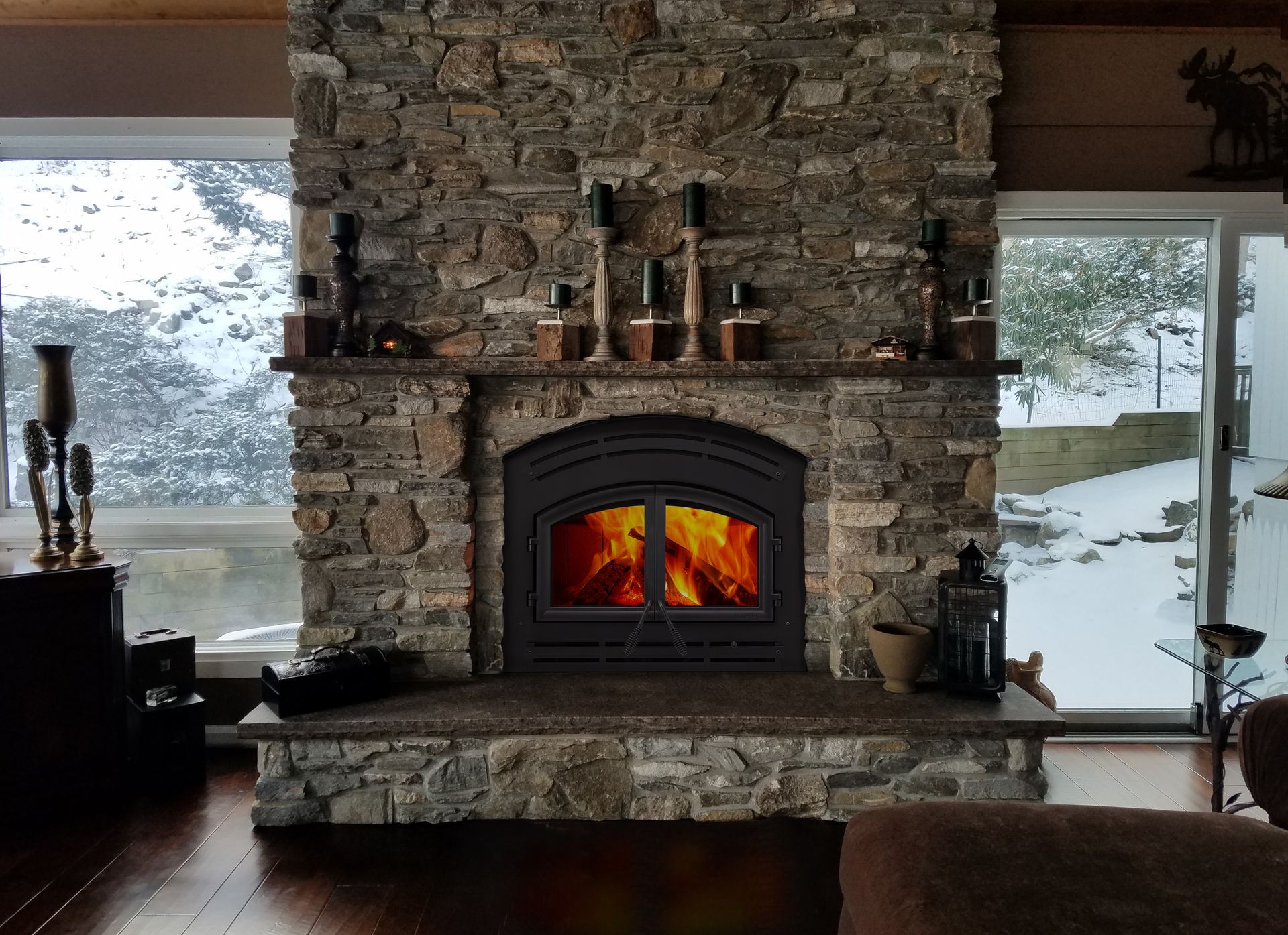 Stone fireplace with a lit fire, large windows, and snowy outdoor view in a cozy room