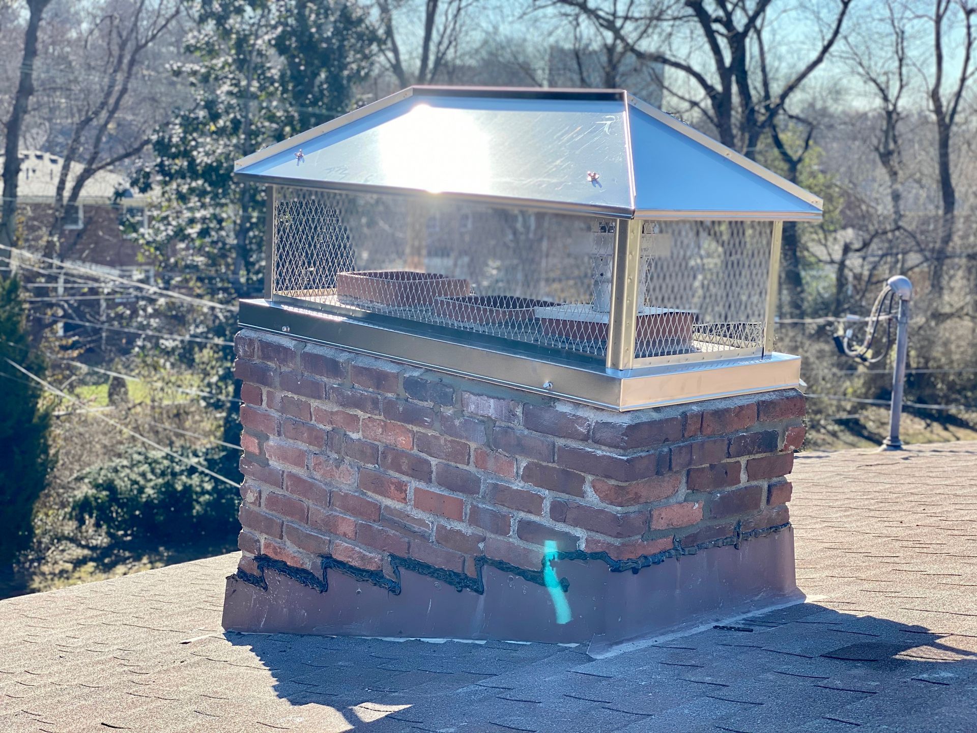Brick chimney with a metal  chimney cap and mesh screen in a wooded outdoor setting