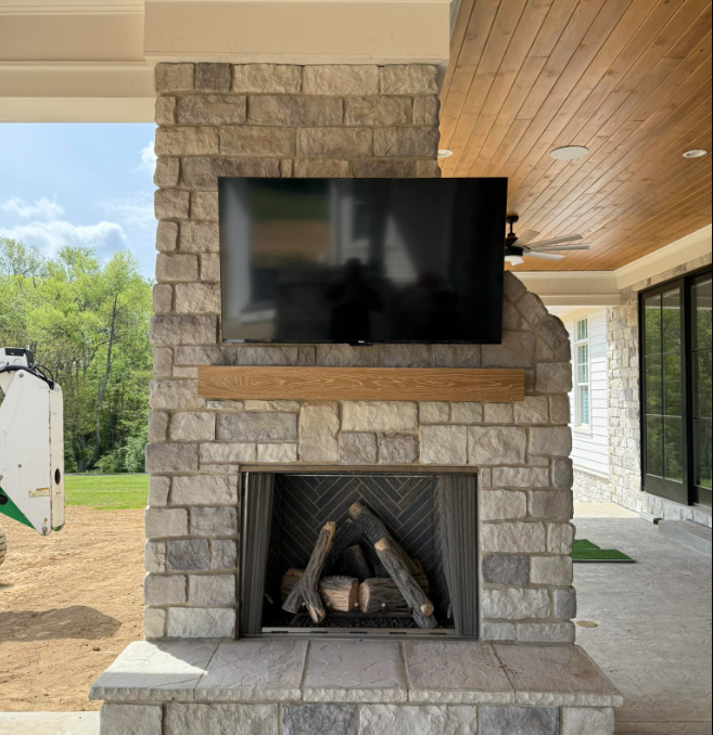 Stone outdoor fireplace with mounted TV under a covered patio, overlooking a grassy yard