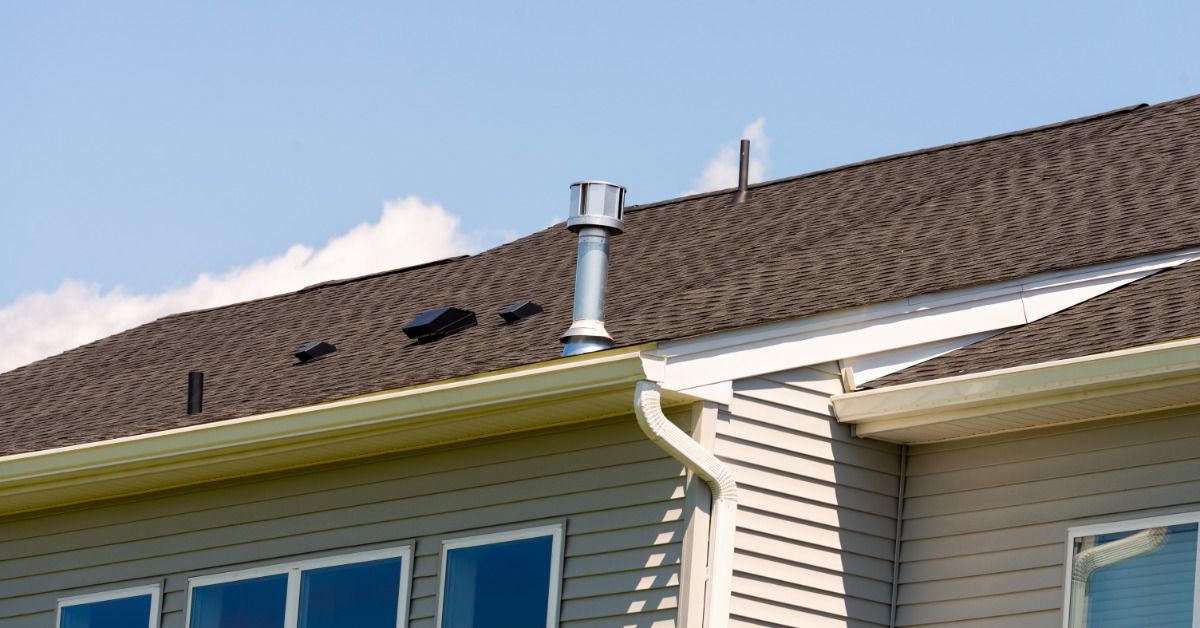 Brown shingle roof with a metal ventilation pipe and small roof vents above a beige-sided home under a clear blue sky.
