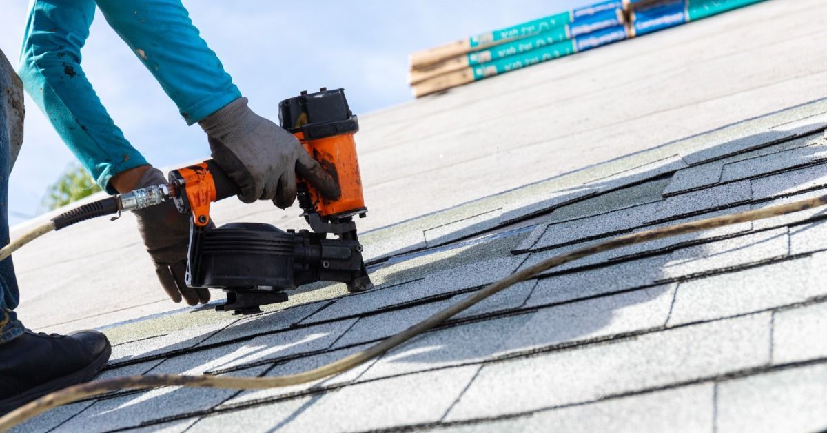 Close-up of roofer using an orange pneumatic nail gun to install gray asphalt shingles on a sloped r