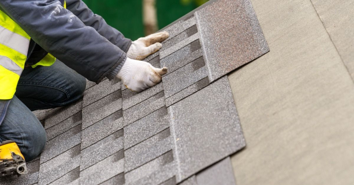 Roofer positions gray asphalt shingles by hand on a sloped roof while wearing safety gear during a roof installation.