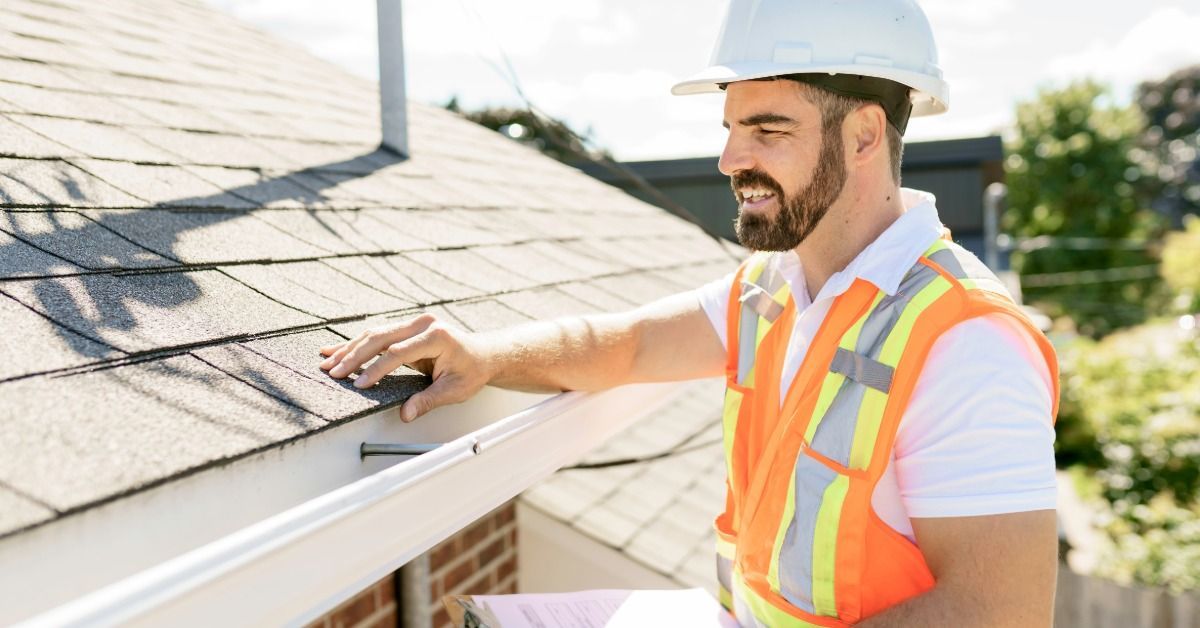 Roofing inspector in a hard hat and safety vest examines asphalt shingles while holding a clipboard 
