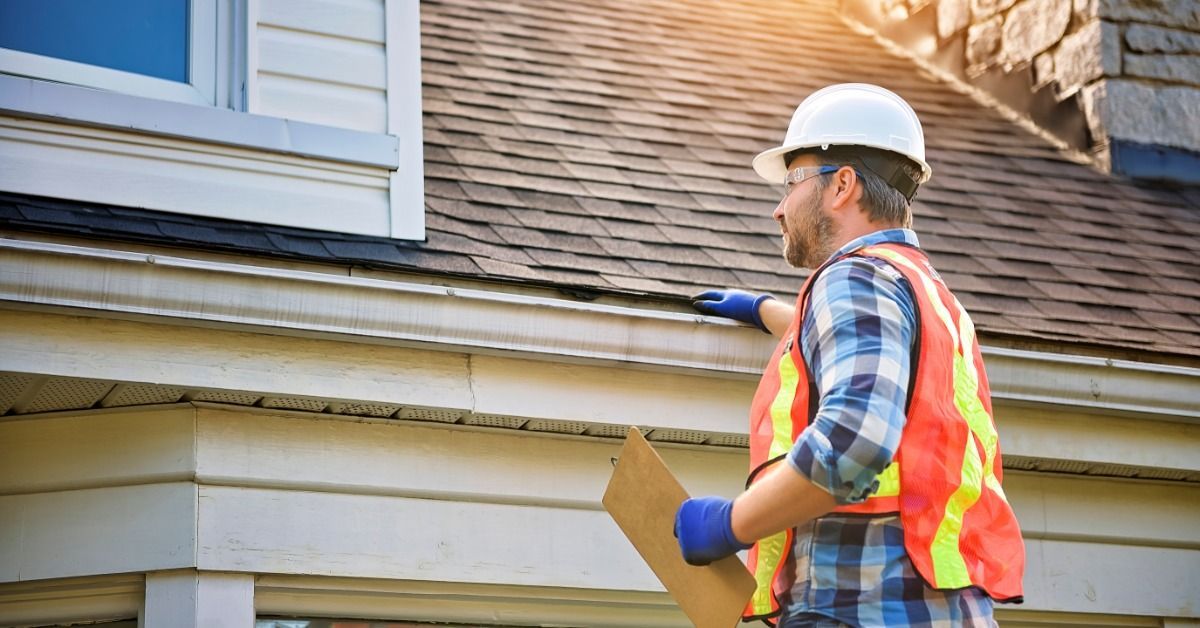 Roof inspector in a hard hat and safety vest examines a home's gutter and shingled roof while holding a clipboard.
