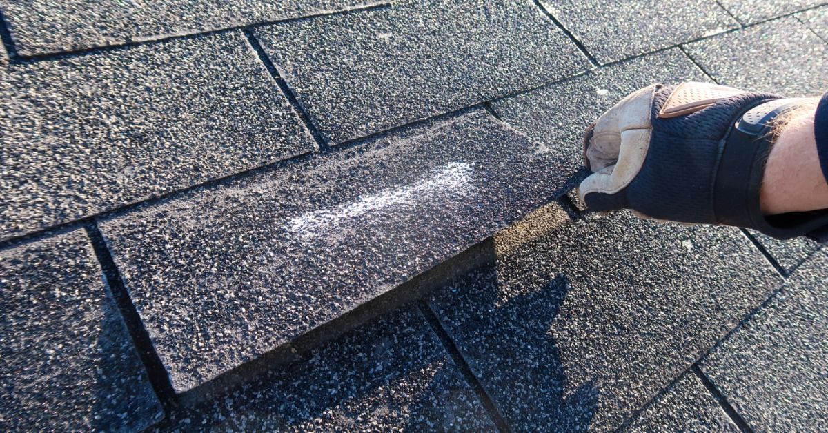 Gloved roofer lifts a damaged asphalt shingle during a close-up inspection of a weathered residential roof.